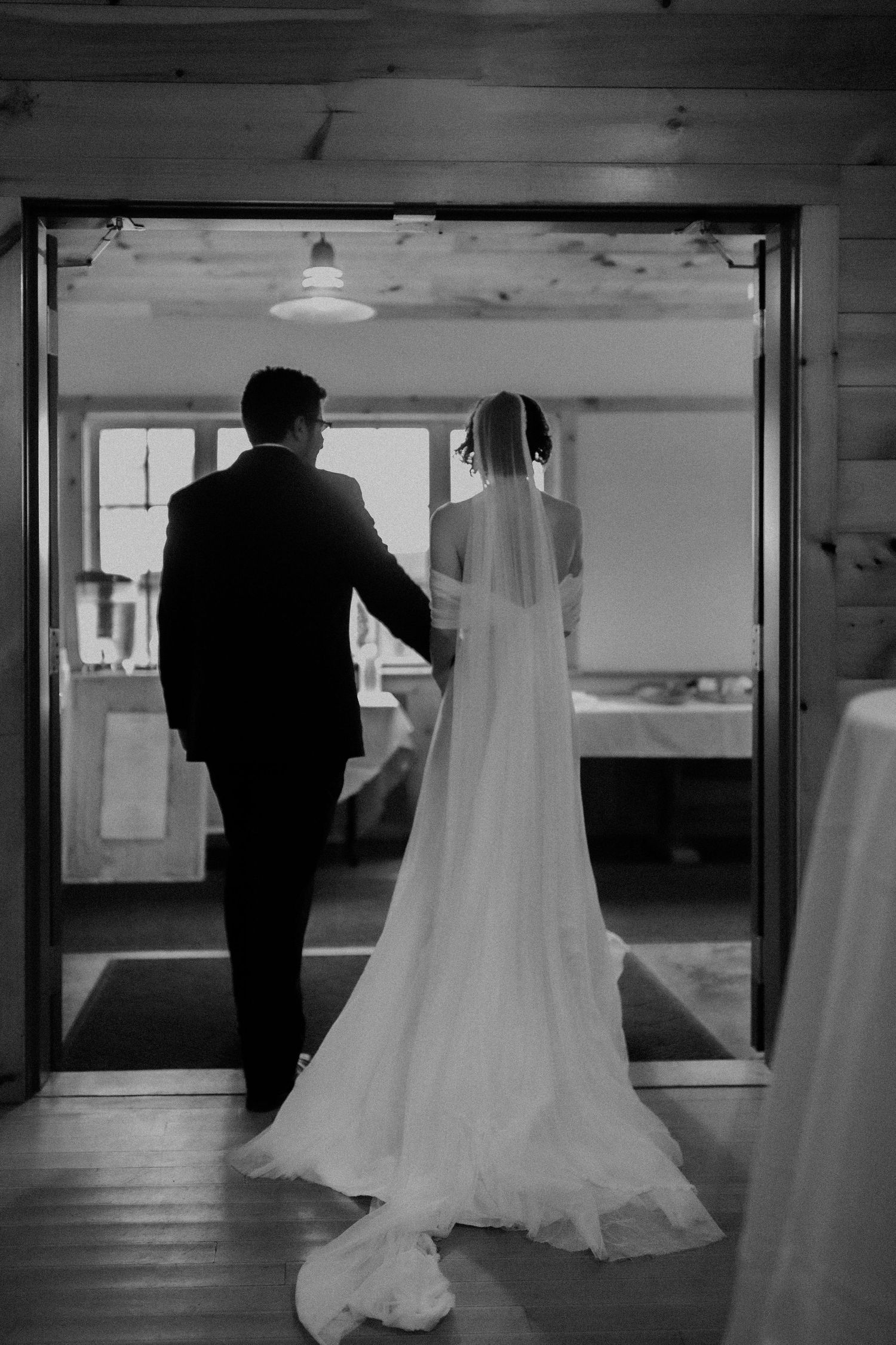 A back view of a couple walking through a doorway in wedding attire with a flowing white dress train.