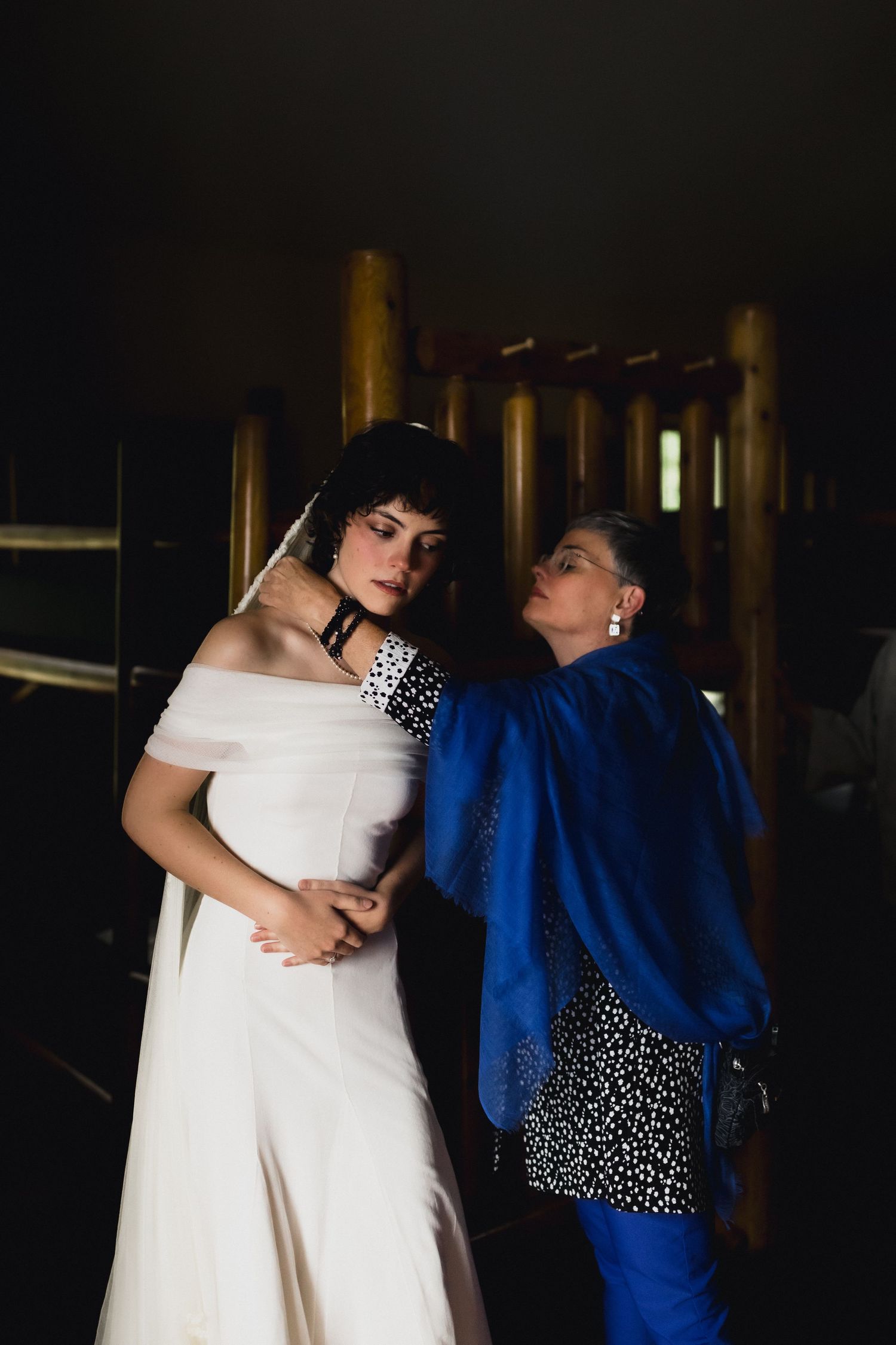 A bride in an off-shoulder white dress poses with someone in blue in a dark interior setting.