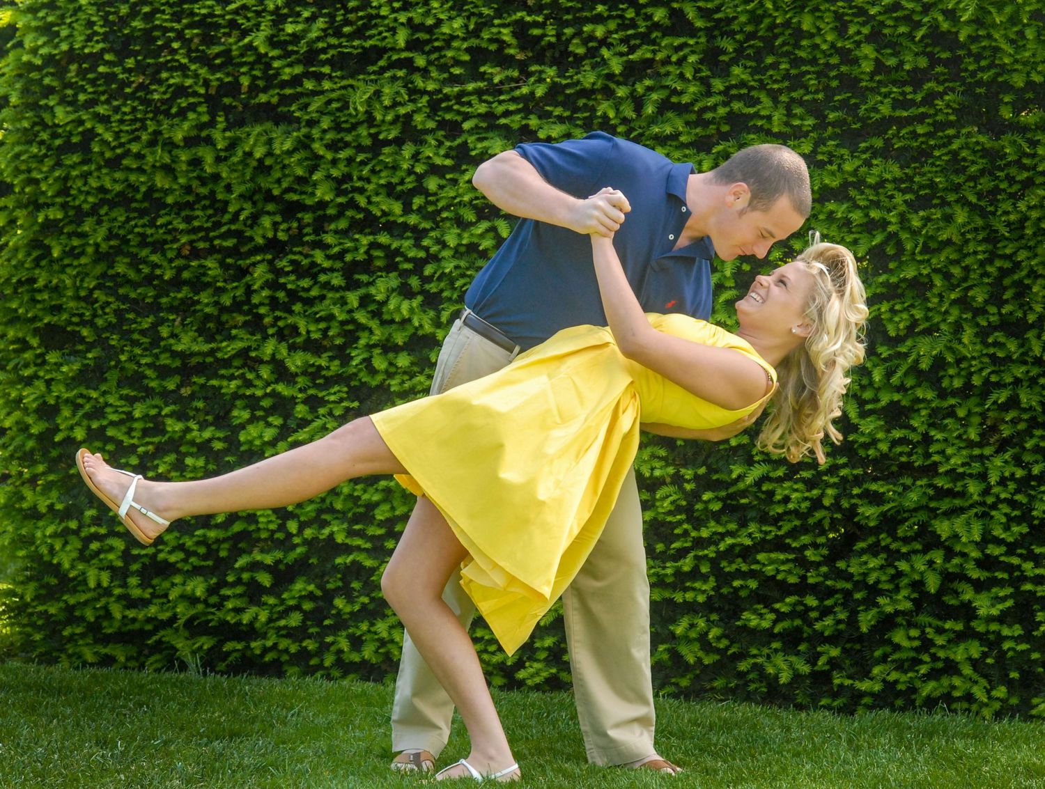 A couple embraces in a romantic dip pose in front of a lush green hedge as the woman in a yellow dress leans back.