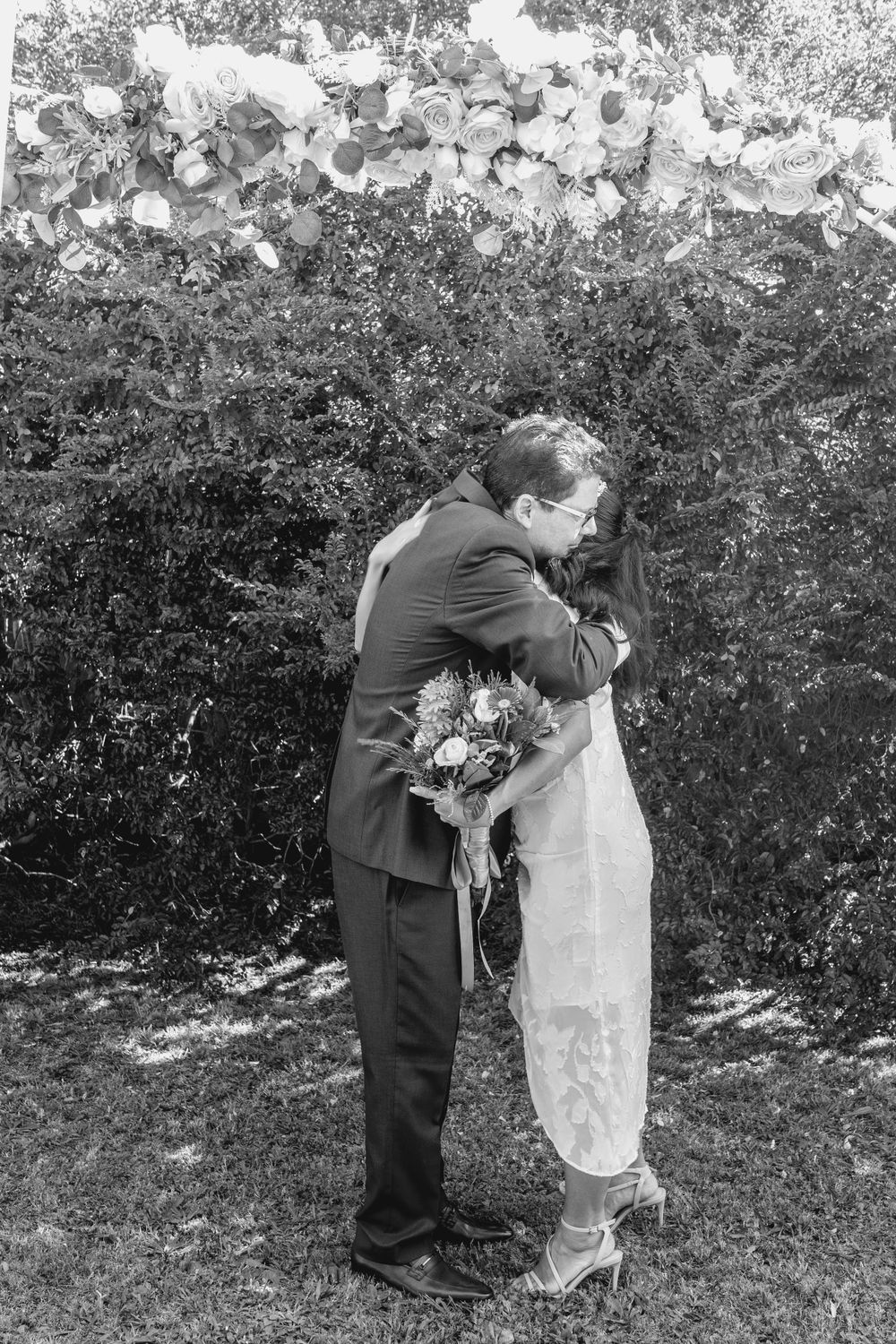 mature asian bride and mature groom embracing under floral arbour after first kiss