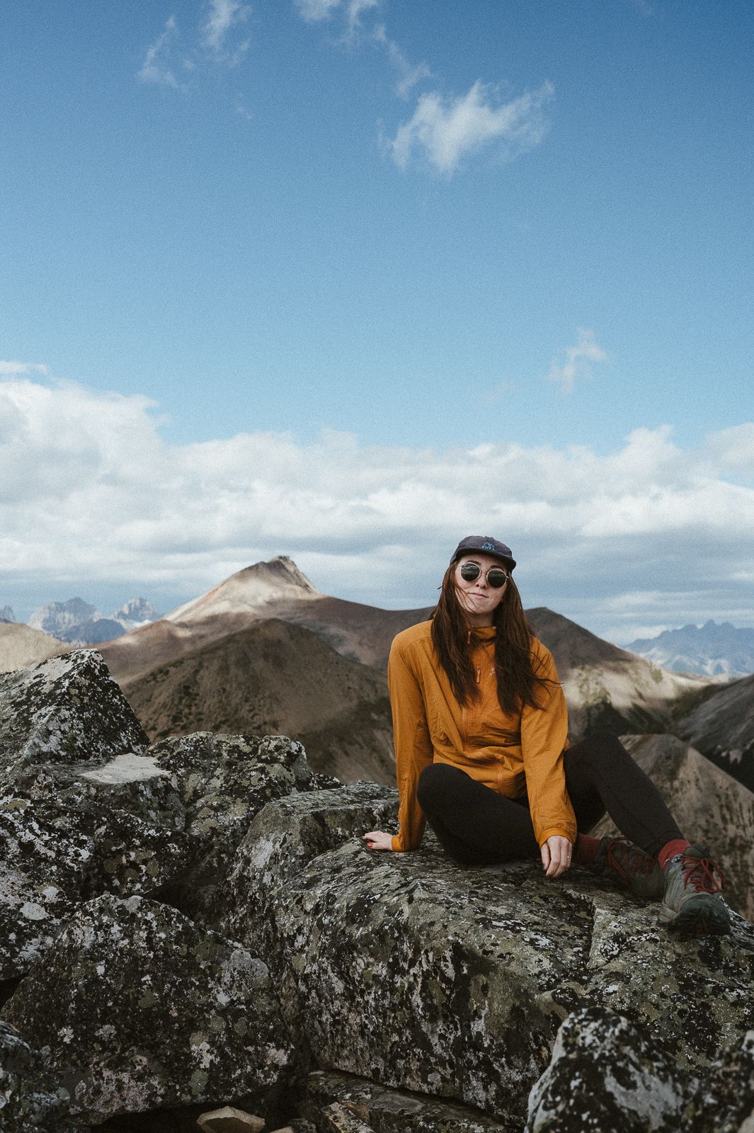 Adventurous hiker in yellow sweater sits atop rocky mountain peak with scenic mountain vista in background.
