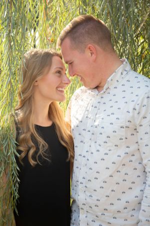 Smiling couple gazes at each other under weeping willow branches in golden sunlight.