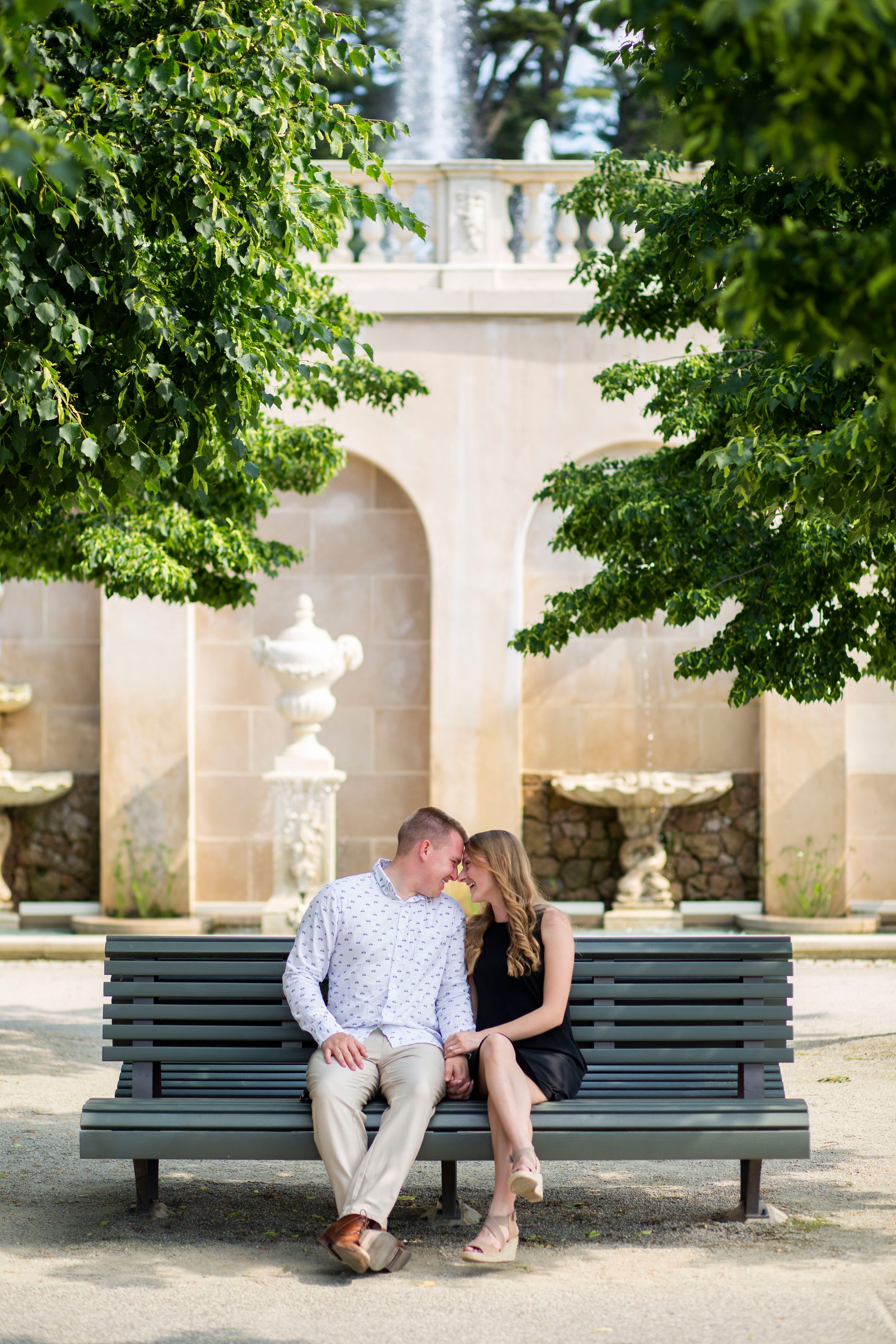Couple sits close on a bench near a fountain surrounded by lush green trees.