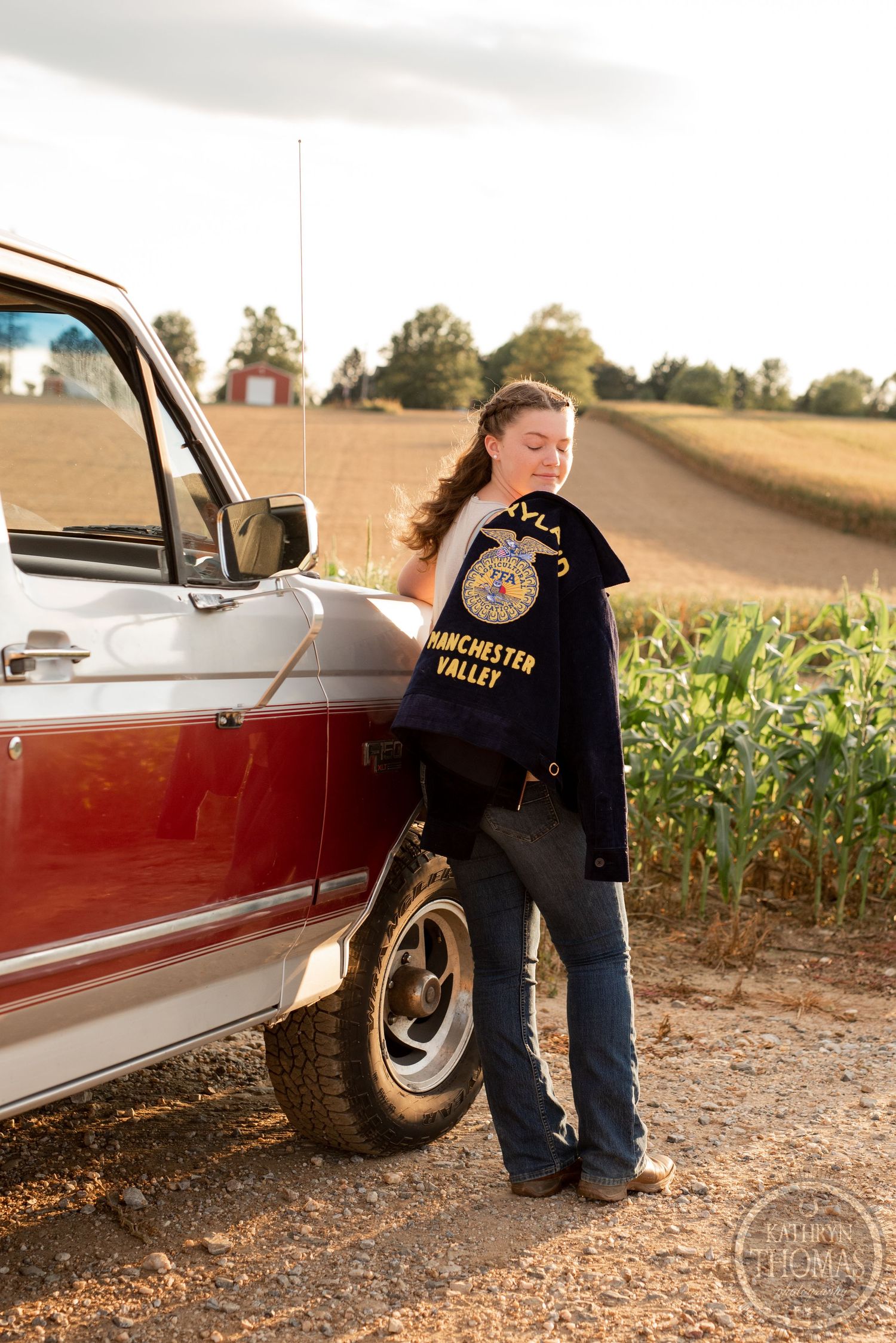 Standing beside a vintage red truck wearing an FFA jacket near a cornfield at sunset.