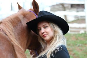 A rider in a black hat and embroidered western wear stands close to a chestnut brown horse outdoors.