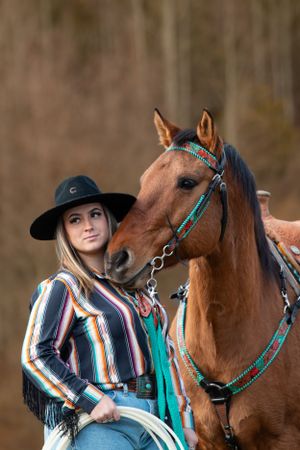 A western-style portrait of a chestnut horse wearing turquoise tack and rider in striped shirt and black hat.