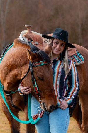 A chestnut horse wearing turquoise tack stands with someone in a striped shirt and black hat in an autumn field.