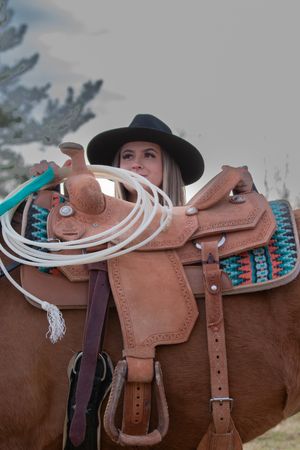 A brown leather western saddle with turquoise accents and white lasso rope rests on a horse.