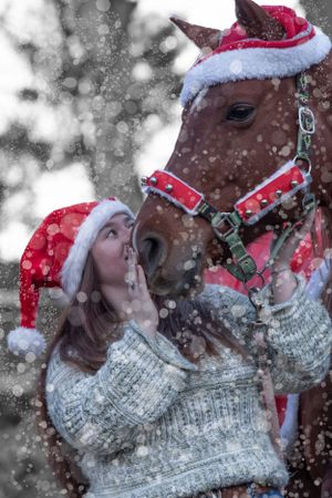 A brown horse wearing a Santa hat stands close to someone in a white sweater and red Santa hat while snow falls around them.