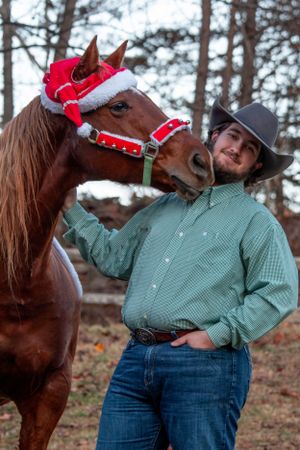 A brown horse wearing a festive Santa hat and halter stands next to a person in a cowboy hat and green shirt outdoors.