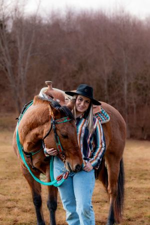 A western-style rider in a black hat stands with their chestnut horse wearing turquoise tack in an autumn field.