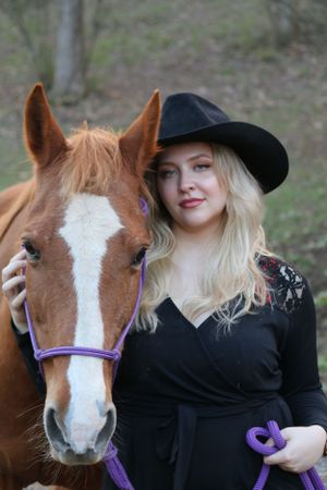 A chestnut horse with white blaze wearing purple halter stands next to someone in black attire and cowboy hat outdoors.