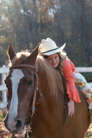 A brown and white horse with leather bridle is being hugged by a rider wearing a white cowboy hat and coral top.