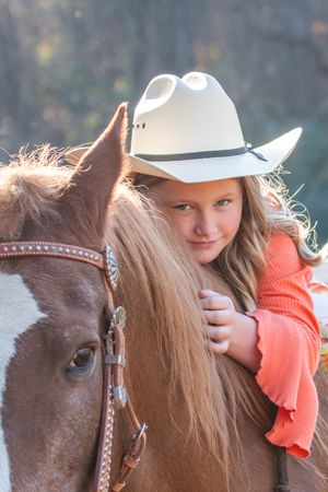 A rider in a white cowboy hat leans against a brown horse with a decorated bridle during a sunny outdoor photo.