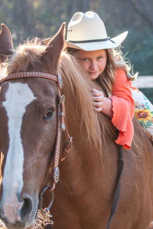 A brown and white horse with decorative bridle is ridden by someone wearing an orange sweater and white cowboy hat.