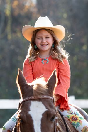 A young rider in a coral sweater and white cowboy hat sits atop a brown and white horse on a fall day.
