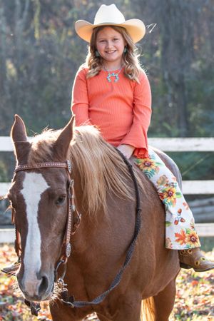 Young rider in orange sweater and cowboy hat sits atop brown and white horse in autumn setting with white fence.
