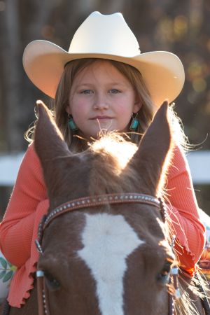 A close-up view between a horse's ears showing a rider in a white cowboy hat wearing a coral colored top during sunset.