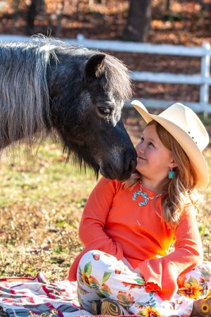 A small black pony and a person in an orange sweater and cowboy hat share a tender moment on a picnic blanket outdoors.