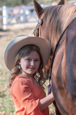 Young equestrian in a cowboy hat and orange sweater smiles while standing next to a brown horse outdoors.