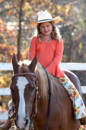 A young rider in a white hat and coral sweater sits atop a brown and white horse on an autumn day.