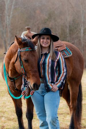A brown horse with turquoise tack and western saddle stands with its rider in a casual country setting on a cloudy day.