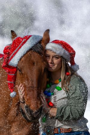 A brown horse decorated in Christmas attire and festive lights stands in the falling snow wearing a Santa hat.