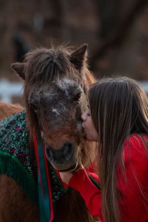 A person in red clothing shares a tender moment with their brown pony wearing a green holiday blanket.