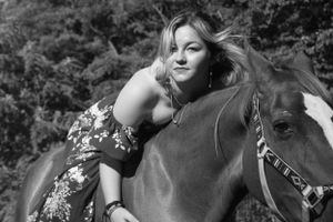 A person in a floral dress leans affectionately against a dark horse outdoors in a black and white portrait.