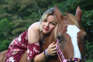 A person in burgundy floral dress embraces a brown and white horse outdoors with green foliage in background.