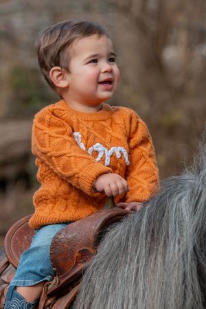 A young child in an orange knit sweater sits smiling in a brown leather horse saddle.