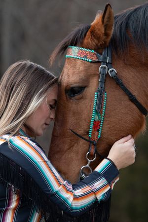 A brown horse wearing turquoise decorative tack is shown being embraced in a tender moment while wearing western style gear.