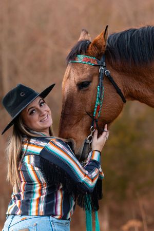A person in western attire and black hat poses with a brown horse wearing turquoise tack against a blurred background.