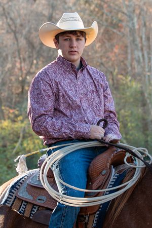 Western cowboy on horseback wearing paisley purple shirt and light cowboy hat holding rope lasso at sunset.