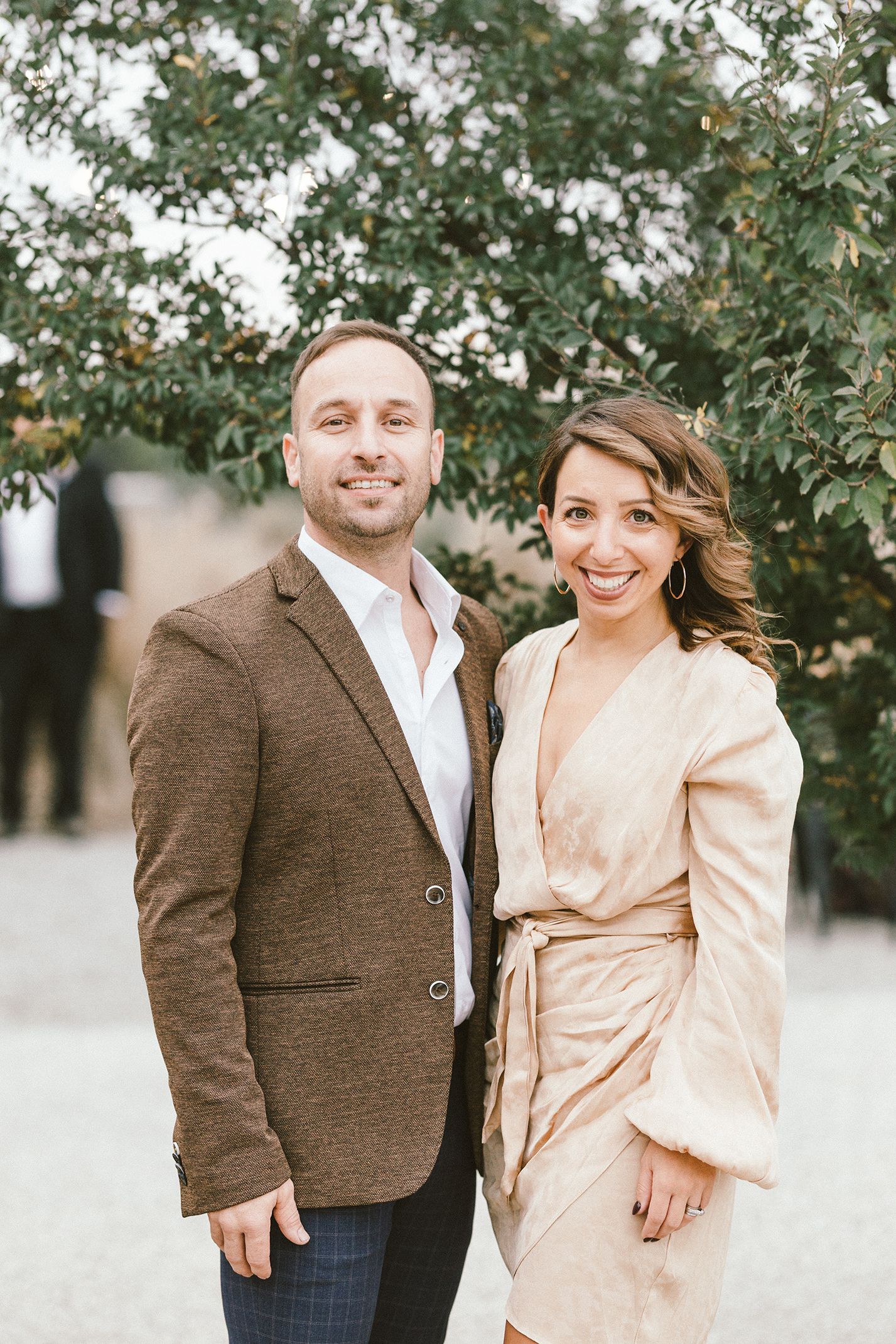 Couple in formal attire posing outdoors under green trees.