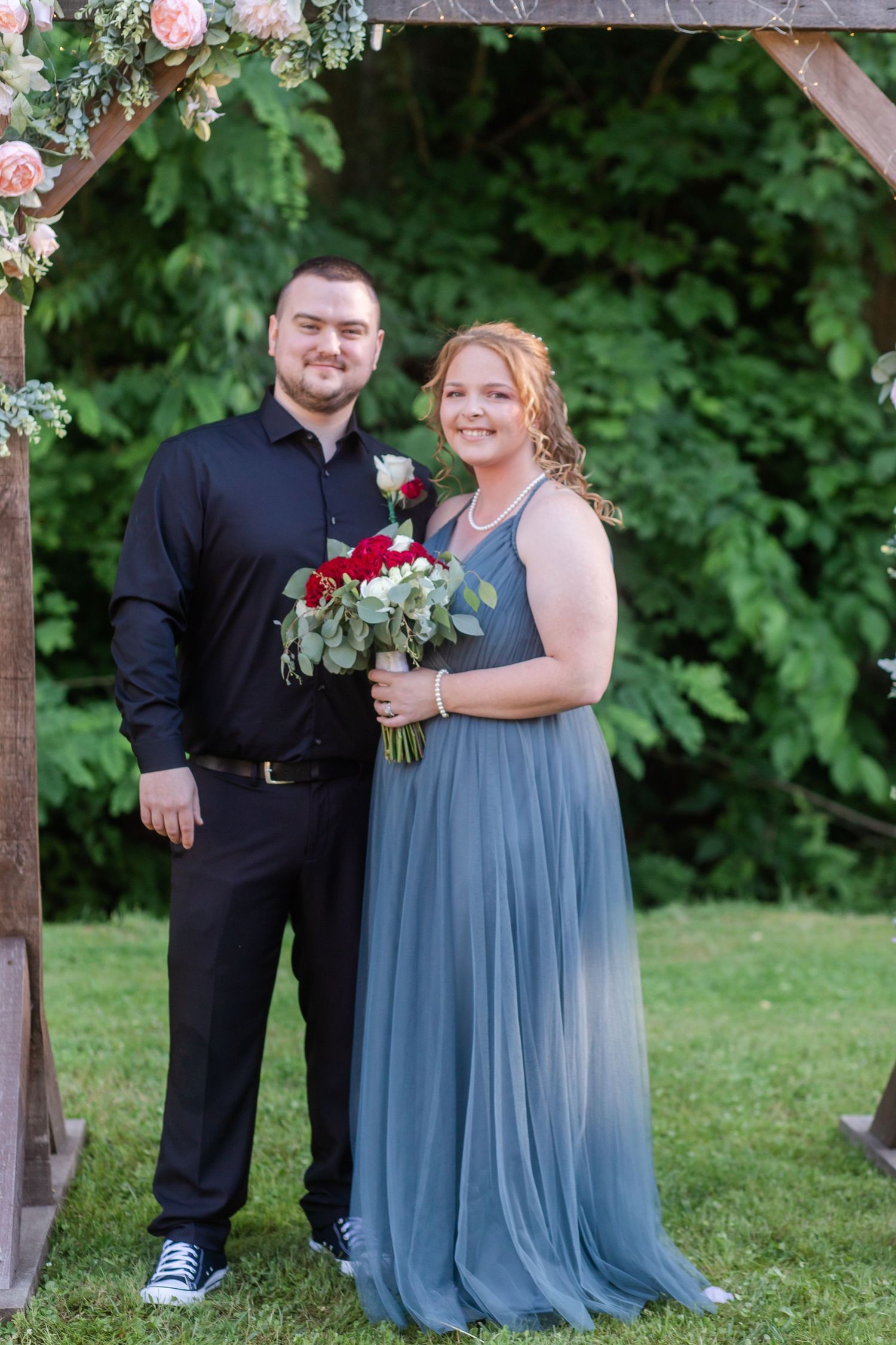 Wedding couple posing under floral arch with dusty blue dress and black suit attire.