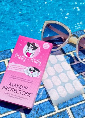 Pink makeup protector packaging and sunglasses displayed on blue tile near swimming pool.