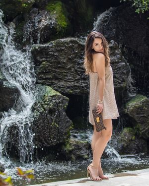 Woman in a beige dress poses near a cascading waterfall in a natural setting. Commercial Photography