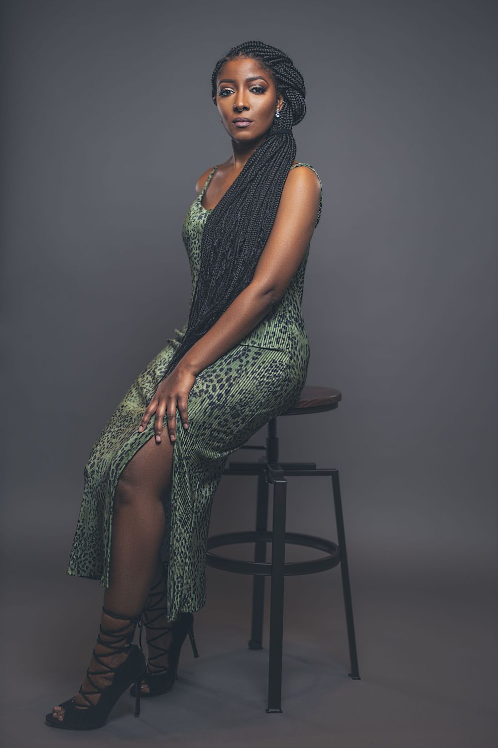 Studio portrait in green sequined dress while seated on black stool against grey background.