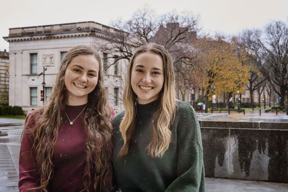 Two friends in burgundy and green sweaters smile together on an autumn day near stone buildings.