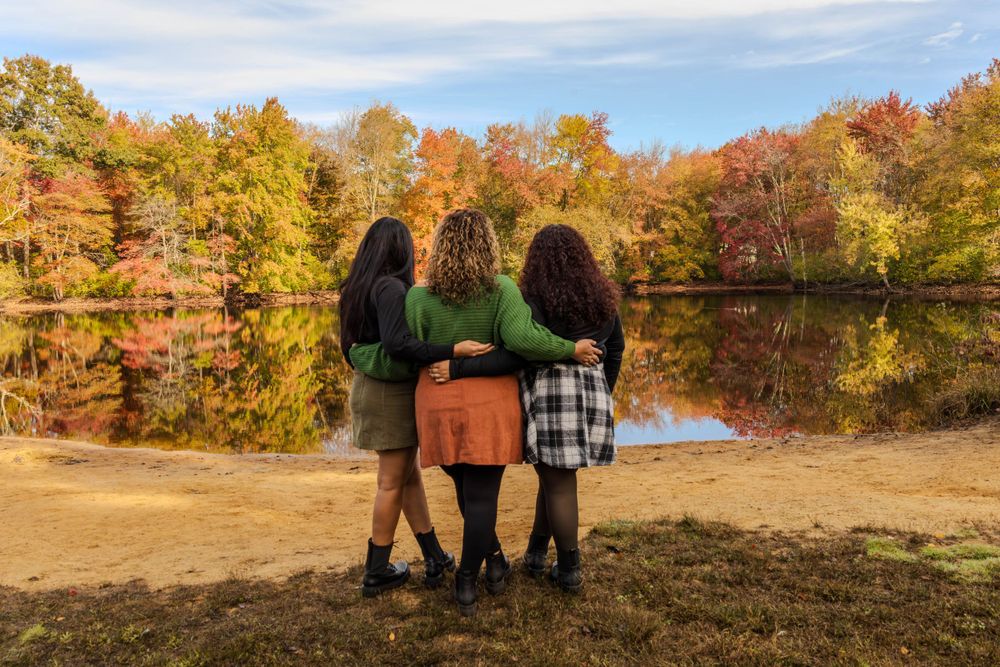 Three friends stand together by a lake during fall with colorful autumn foliage reflecting in water.