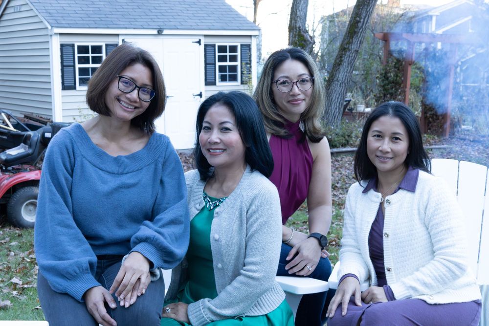 Four women friends sit together outdoors wearing casual sweaters and smiling at camera.