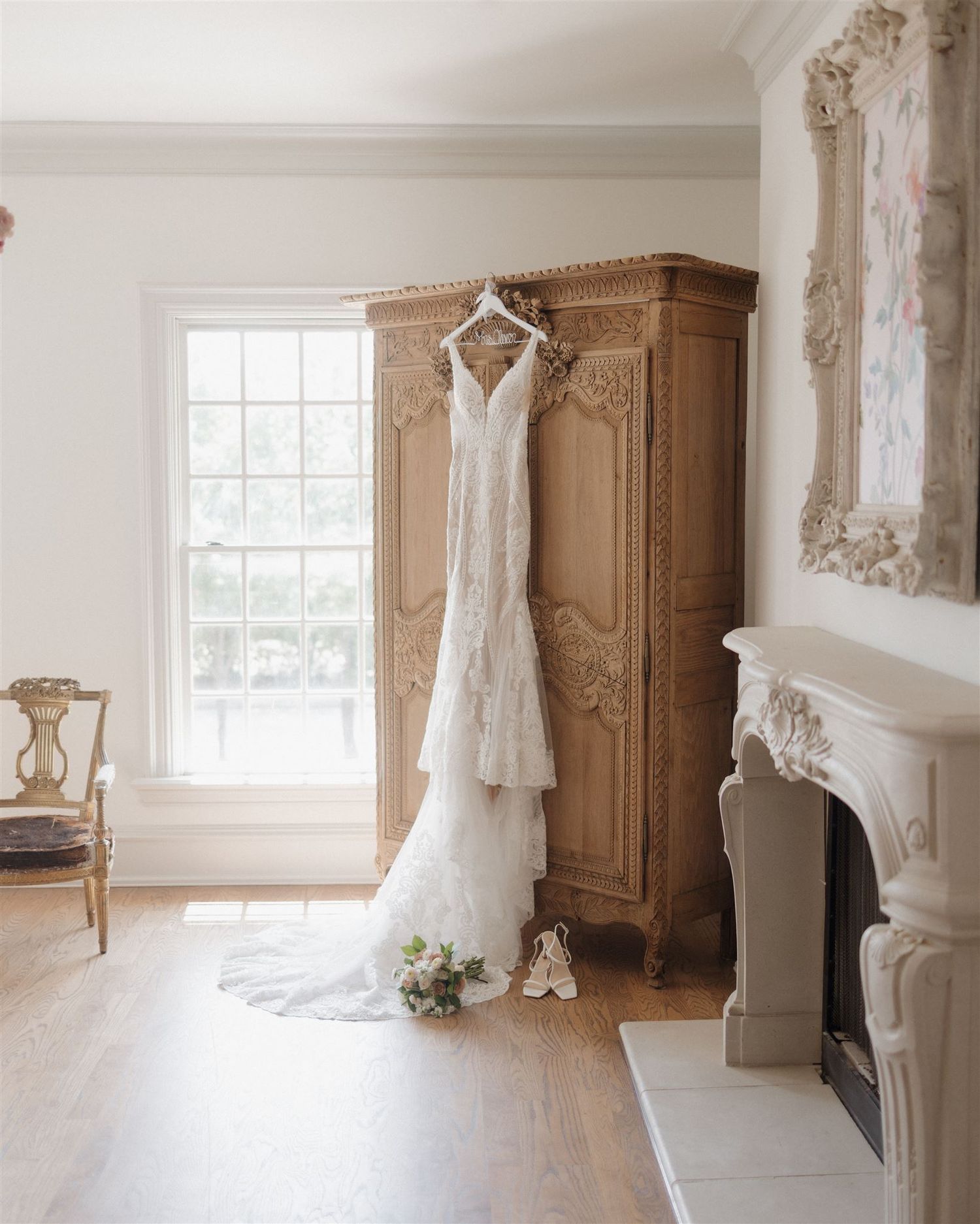 Wedding dress displayed on vintage armoire with bouquet and shoes in elegant white room.