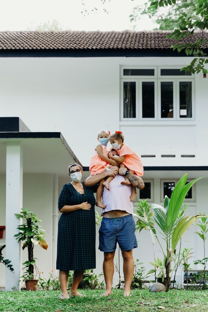 A Dad is holding two daughters who are covering his face, while the mother stands by