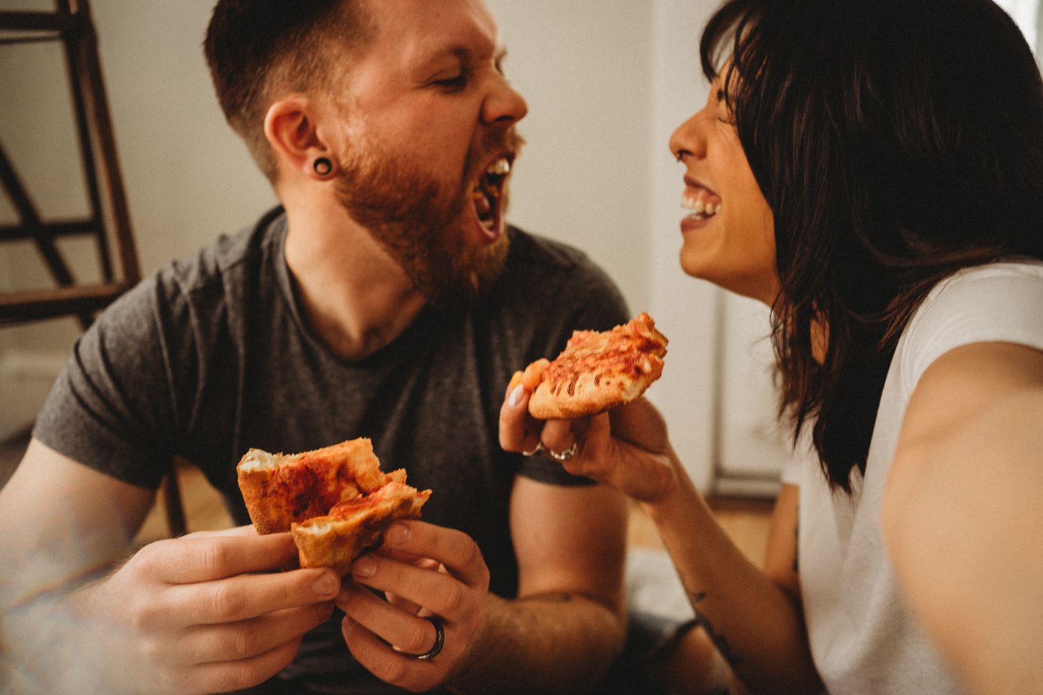 Couple laughing while sharing slices of pizza together in a candid moment.