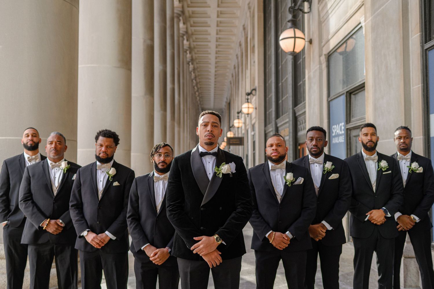 Groomsmen in formal attire pose together under colonnade for wedding photo.