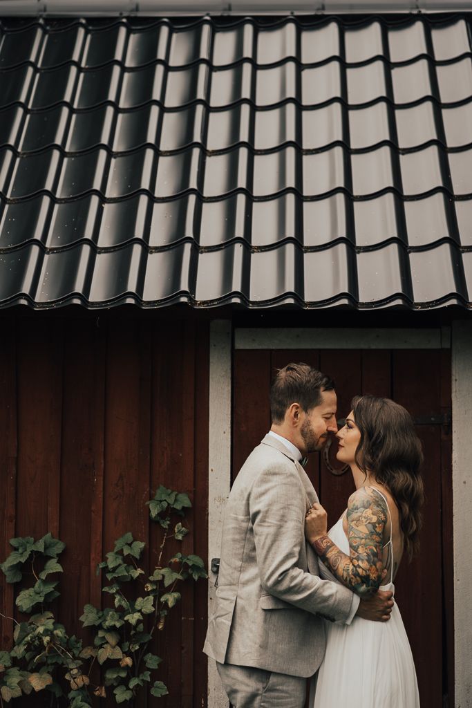 Brudpar myser upp framför en faluröd bod. Bröllopsfotografering i Eskilstuna.