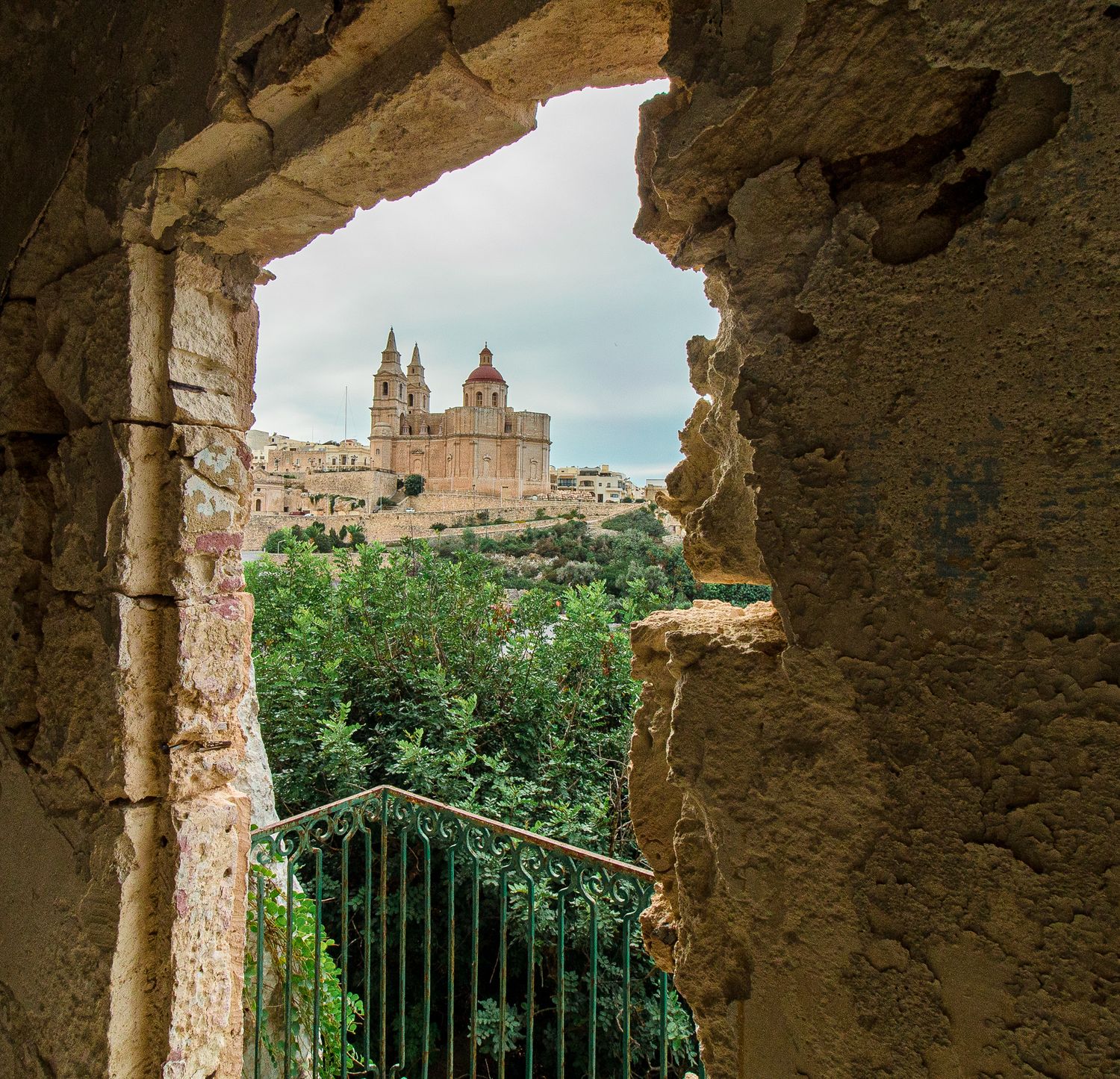 View through broken stone wall opening of historic Malta church and village surrounded by lush green trees.