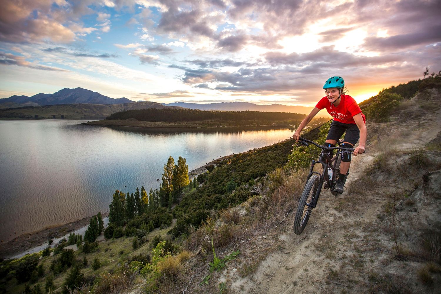 Mountain biker riding a steep dirt trail at sunset with a scenic lake and cloudy sky behind.