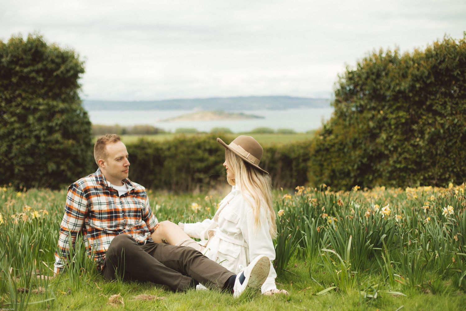 A couple sits together in a blooming daffodil field overlooking coastal views on a sunny spring day.